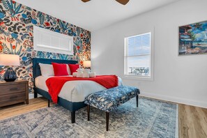 Une chambre à coucher avec un papier peint à motifs floraux, un lit avec une literie rouge et blanche, un banc, une table de chevet et une fenêtre avec des stores.