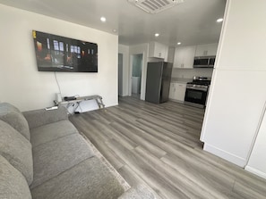 Ein modernes Wohnzimmer mit grauem Sofa, einem flachbildfernseher an der Wand und einer Küchenzeile mit weißen Schränken und Edelstahlgeräten.