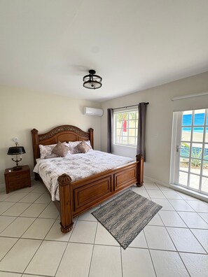 Ein Schlafzimmer mit einem hölzernen Bett, einem Nachttisch mit Lampe, einem Fenster mit Vorhängen und Blick auf den Strand.