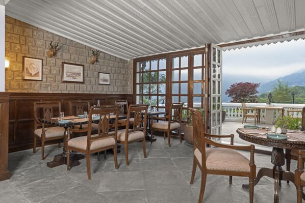 A restaurant with a stone wall, wooden chairs, a bar, and large windows offering a mountain view. - Brij Anayra Dharamshala (Dharamshala)