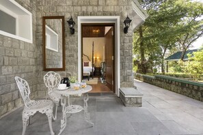 A stone building with a patio, white ornate chairs, a small round table, and a doorway leading to a room with a bed and nightstand. - Brij Anayra Dharamshala (Dharamshala)