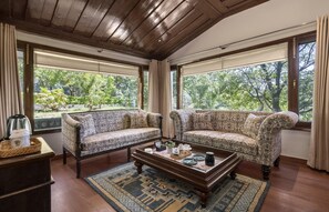 A room with two patterned sofas, a wooden coffee table, and a view of greenery through large windows. - Brij Anayra Dharamshala (Dharamshala)
