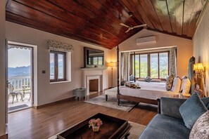 A spacious room with a wooden ceiling, a fireplace, a bed with pillows, a sofa, and a view of mountains through large windows. - Brij Anayra Dharamshala (Dharamshala)