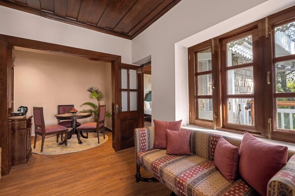 A room with a wooden floor, a sofa with patterned cushions, a dining table with chairs, and a large window with multiple panes. - Brij Anayra Dharamshala (Dharamshala)