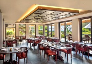 Un'ampia sala da pranzo con diversi tavoli e sedie, grandi finestre che offrono vista sul verde e un moderno design del soffitto.