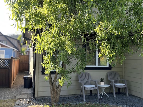 Un árbol con hojas verdes frente a una casa, dos sillas y una pequeña mesa.