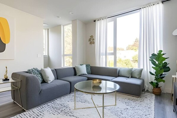 A modern living room with a sectional sofa, a glass coffee table, and a large window with curtains.