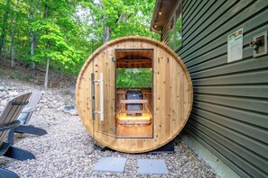 Estructura de sauna de madera con un banco en su interior, rodeada de grava y árboles.