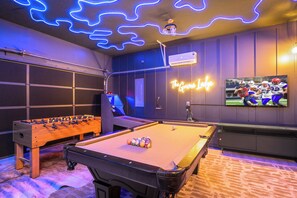 Una habitación con mesa de billar, mesa de futbolín y un televisor mostrando un partido deportivo.