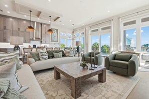 Ein geräumiges Wohnzimmer mit einer großen Couch, einem Couchtisch und einem grünen Sessel. Es gibt eine Küchenzeile mit Schränken und einem Esstisch.