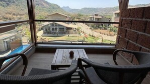 A covered balcony with wicker chairs, a wooden table, and a view of a mountainous landscape. - Mastiff Grand La Villae Resort, Lonavala (Mawal)