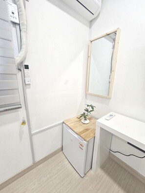 Et lille, rent værelse med hvid væg, træbordplade, lille køleskab, spejl og en aircondition.