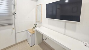 Ein kleiner, sauberer Raum mit weißer Wand, einem angebrachten Fernseher, einer kleinen Kühlbox und einem Holzschrank.