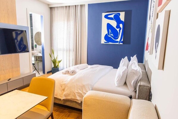 Habitación de hotel con cama, escritorio, silla, televisor y ventana con cortinas.