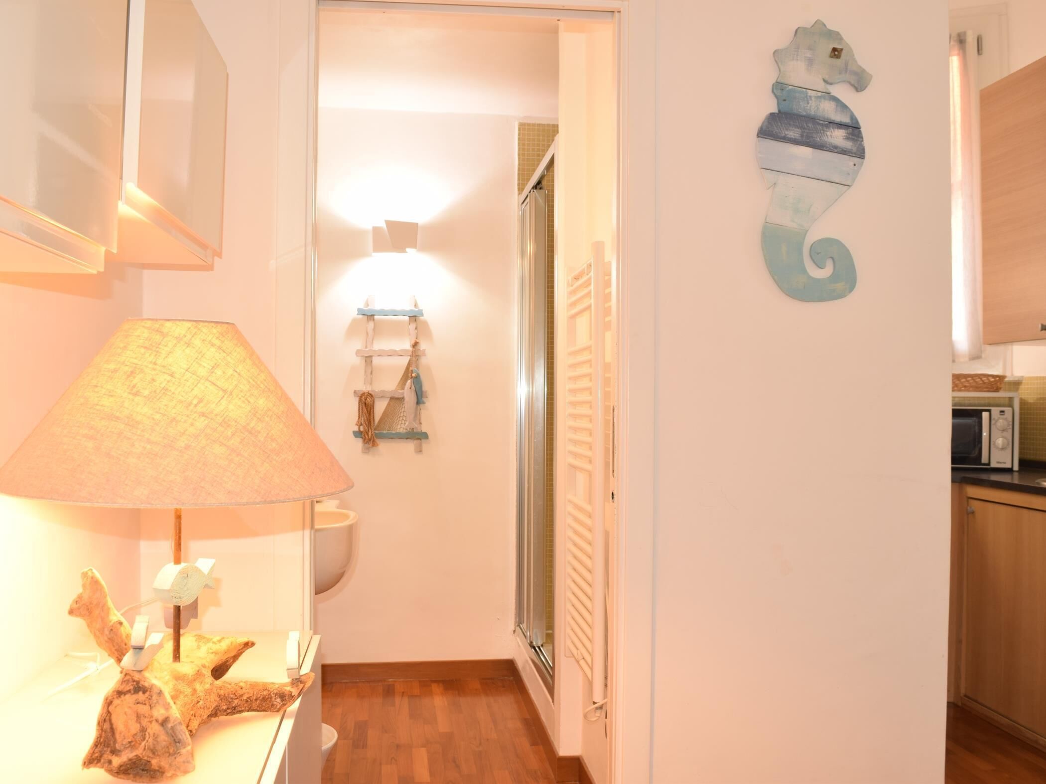 A hallway with a lamp, a seahorse wall decoration, a wooden floor, and a microwave in the kitchen.