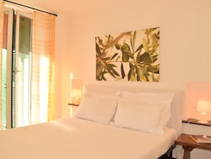 Ein Hotelzimmer mit Bett, Nachttisch, Lampe, einem Fenster mit Vorhängen und einem an der Wand hängenden Bild.