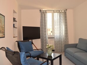 Ein Wohnzimmer mit einer Couch, einem Sessel, einem Fernseher, einem Tisch mit einer Vase und gemusterten Vorhängen.