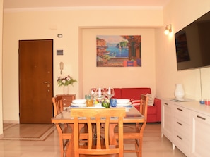 A dining area with a wooden table and chairs, a red sofa, a painting on the wall, and a TV mounted above the sofa. - Rental unit in Rapallo - 2 bedrooms 1 bathroom air conditioned (RAPALLO)