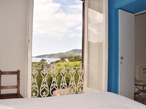 Ein Schlafzimmer mit Blick auf eine Küstenlandschaft, ein Balkon mit schmiedeeisernem Geländer und eine blaue Tür.