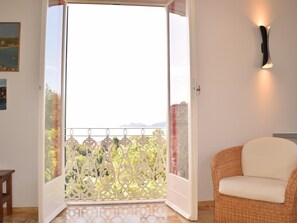 Eine offene Schiebetür führt zu einem Balkon mit Blick auf Grünflächen und Gebäude, ein Korbstuhl und eine Wandleuchte sind vorhanden.
