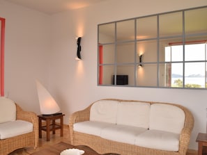 Ein Zimmer mit einem Korbsessel, einem kleinen Tisch, einer Lampe und einem großen Fenster mit Blick.