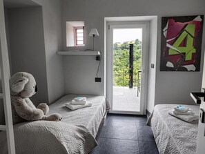 Ein Schlafzimmer mit zwei Betten, einem Fenster, einer zur Außenwelt führenden Tür, einer Lampe und einer Wand mit einem geometrischen Gemälde.
