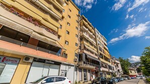 A multi-story building with balconies, a white car parked in front, and various storefronts including a shoe store and a clothing store. - Stone City Stay Central Gjirokast r Apartment (Gjirokastër)