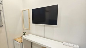 Ein Zimmer mit weißem Schreibtisch, einem kleinen Hocker, einem an der Wand befestigten Fernseher, einem Minikühlschrank und einer Klimaanlage.