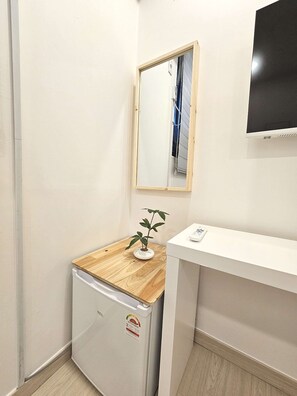 Et lille tekøkken med et kompakt køleskab, en træbordplade, en potteplante, et spejl og et hvidt skrivebord.