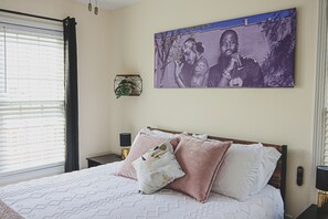 Ein Schlafzimmer mit einem Bett, einem Nachttisch, einem Wandbild, einem Fenster mit Jalousien und einer Pflanze auf dem Nachttisch.
