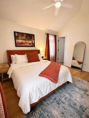 Ein Schlafzimmer mit Bett, Deckenventilator, Nachttisch, Lampe, Gemälde, Spiegel und einer Tür.