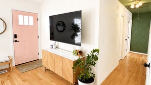 Een hal met houten vloer, een witte deur, een aan de muur bevestigde televisie, een potplant en een groene wand.