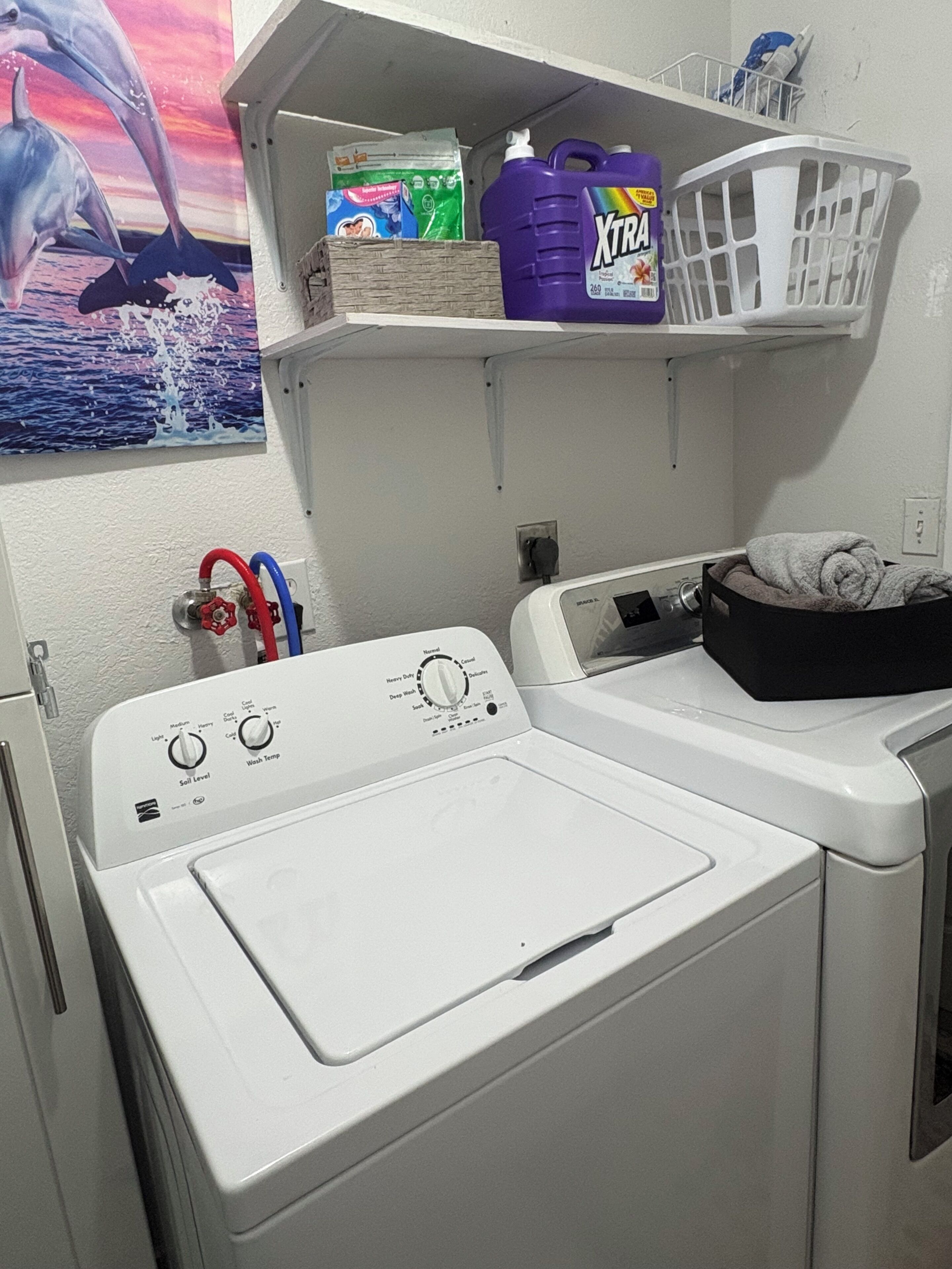 Un espace buanderie comprenant un lave-linge et un sèche-linge à chargement frontal, une étagère avec des produits de nettoyage et une prise de courant fixée au mur.
