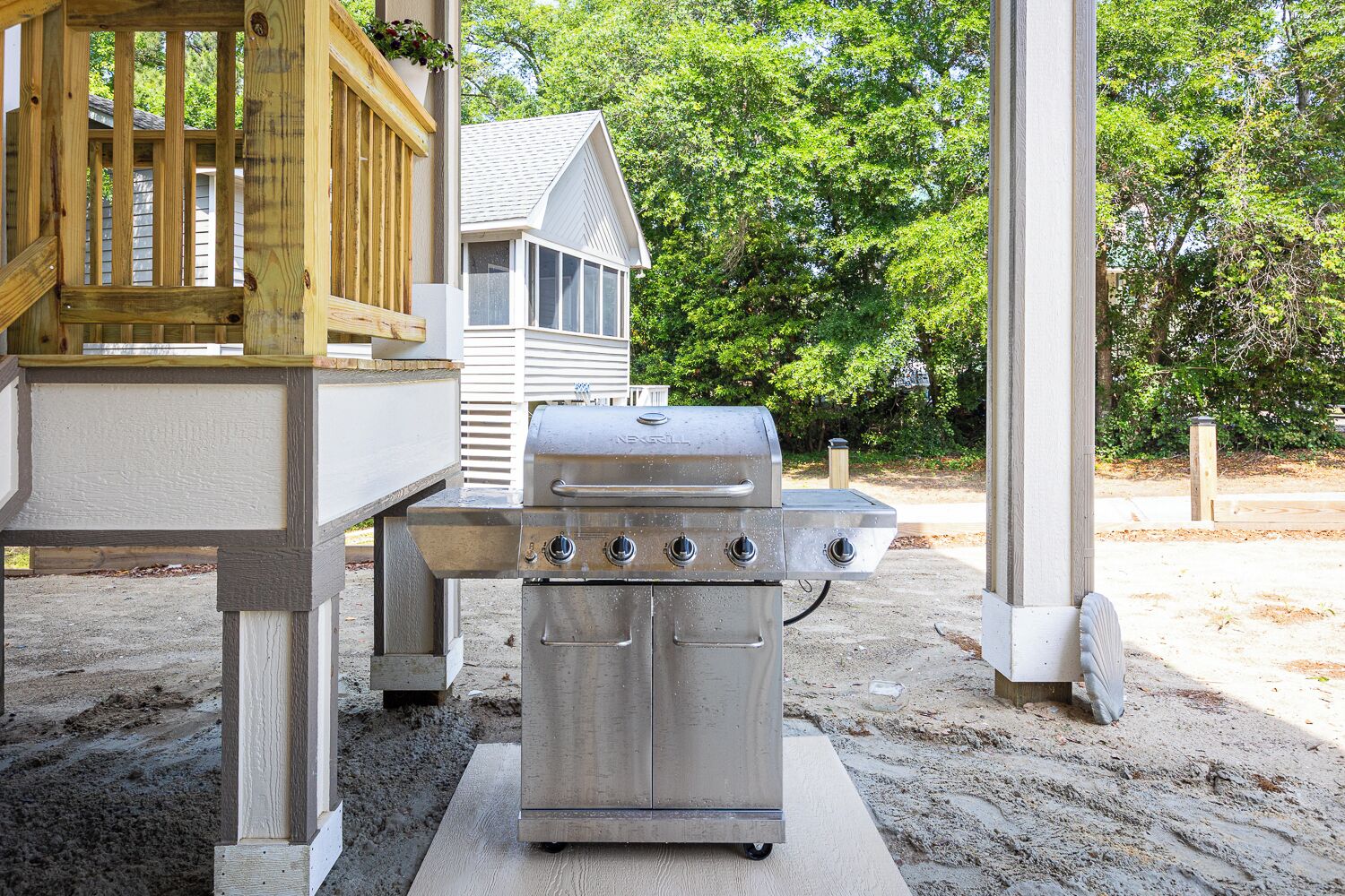 Een draagbare grill van roestvrij staal met vier branders, geplaatst op een betonnen plaat in een achtertuin met een houten terras en een wit huis op de achtergrond.