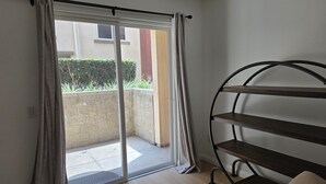 Una habitación con puerta corrediza de vidrio que da a un balcón, una estantería metálica y un sillón tapizado.