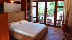 Ein Schlafzimmer mit Bett, Holz-Kopfteil, einem Fenster mit Blick ins Grüne und einem Balkon mit Tisch und Stühlen.