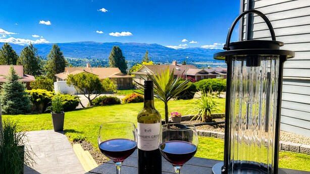 A patio with a bottle of wine and two glasses on a table, overlooking a residential area with houses and mountains. - Spacious Modern King Suite • Patio BBQ • Views (Kelowna)