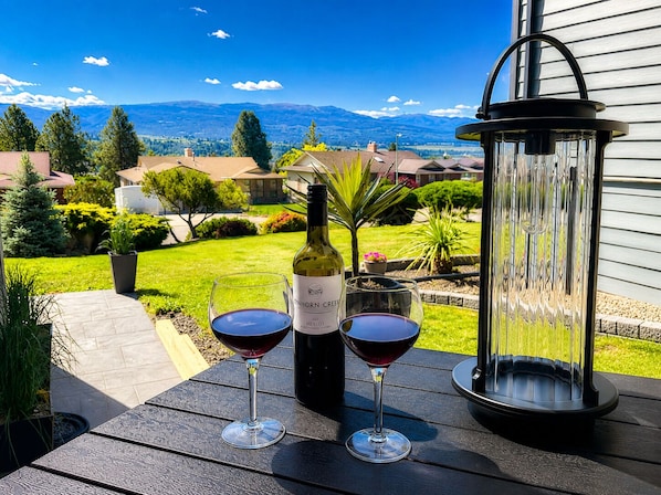 Un patio con una botella de vino y dos copas sobre una mesa, con vistas a una zona residencial con casas y montañas.