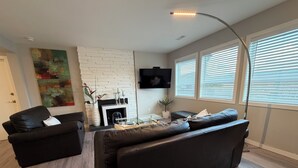 A modern living room with a fireplace, black leather sofas, a flat-screen TV, and a large window with blinds. - Spacious Modern King Suite • Patio BBQ • Views (Kelowna)