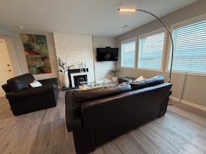 Una sala de estar moderna con chimenea, sofás de cuero negro, un televisor de pantalla plana y un amplio ventanal con persianas.