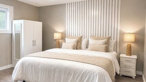 A bedroom with a large bed, two bedside tables with lamps, a wardrobe, and a window with blinds. - Spacious Modern King Suite • Patio BBQ • Views (Kelowna)