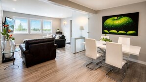 A modern living room with a black leather sofa, a dining table with white chairs, and a large green apple artwork on the wall. - Spacious Modern King Suite • Patio BBQ • Views (Kelowna)