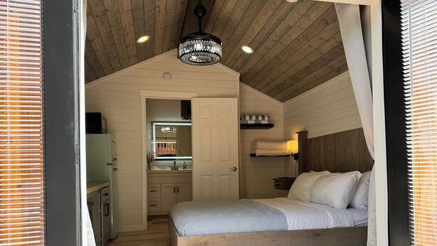 A bedroom with a wooden ceiling, a bed, a nightstand, a mirror, and a small bathroom with a sink and towel rack. - Brand NEW Bungalow in Eatonville - Near Mt. Rainier (Eatonville)