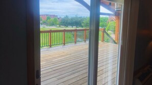 On aperçoit, à travers une porte vitrée, une terrasse en bois avec une rambarde en métal, un bâtiment en briques et, au fond, une zone herbeuse.
