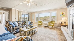 A living room with a patterned sofa, a coffee table, wicker chairs, a ceiling fan, and large windows. - Direct Ocean Front 3BR w/ Pool (Nags Head)