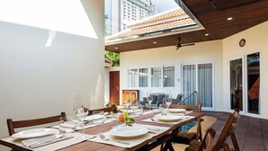 A dining area with a long table set for four, surrounded by chairs, with a view of a building and a covered patio. - Luxury 4BR Pool Villa Pratamnak Soi5 - 50m to Beach (Pattaya)