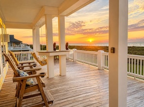 A wooden deck with a view of the sunset over the ocean, a table, and two chairs. - Beautiful 4BR Beachfront | Elevator + Game Room (Port Bolivar)