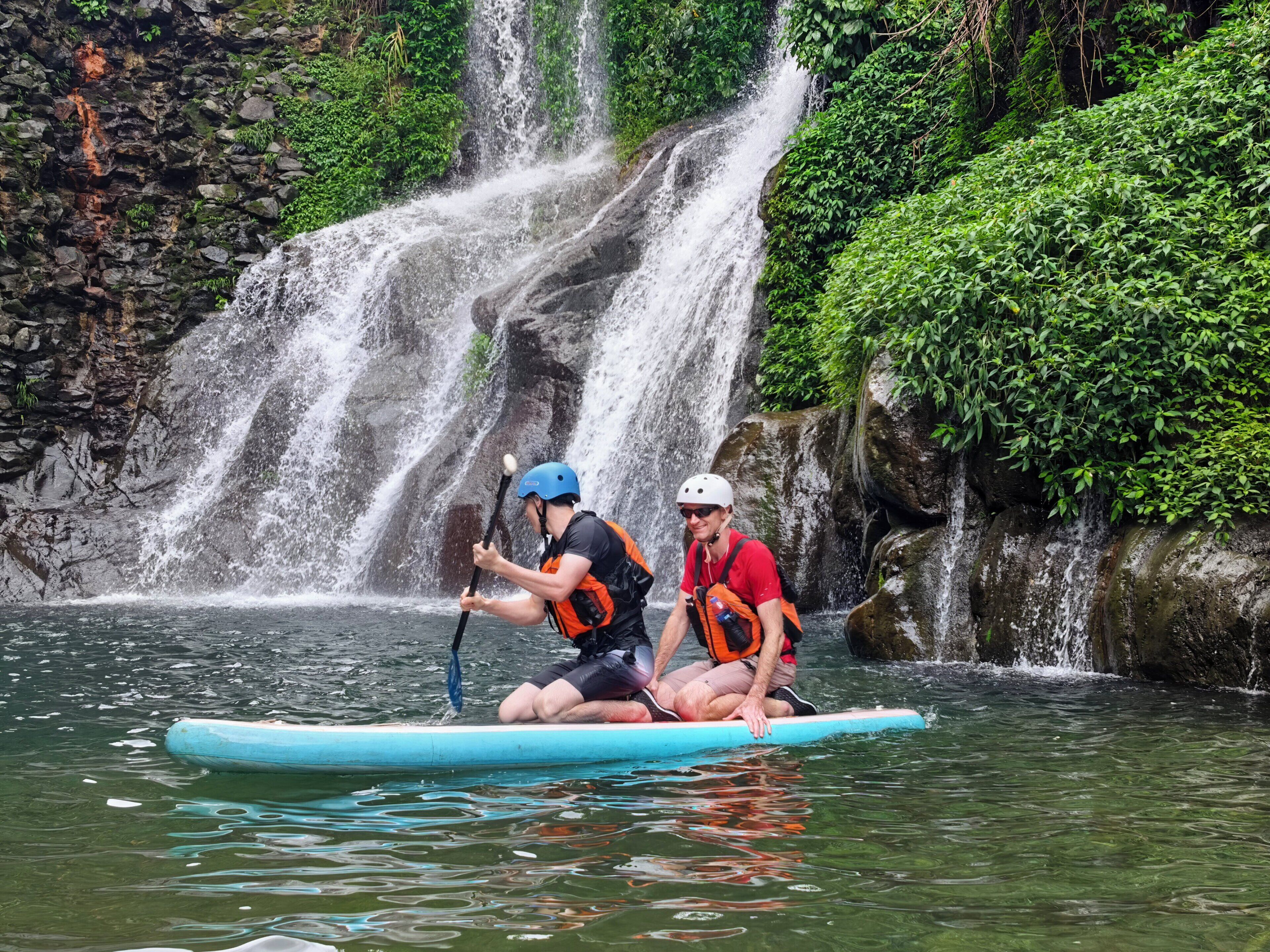 Two people on a blue raft in a river with a waterfall in the background.