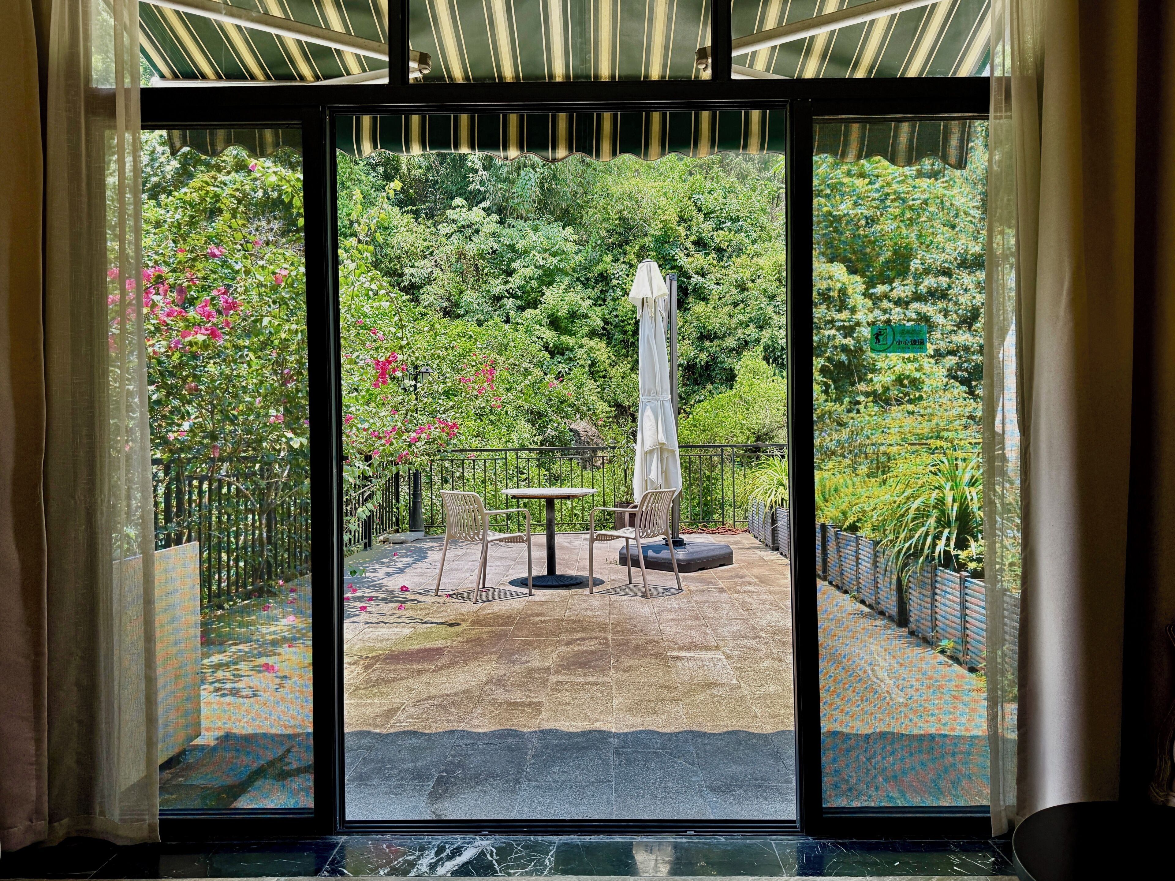 A view through a glass door into an outdoor patio area with a table and chairs, surrounded by lush greenery and a metal fence.