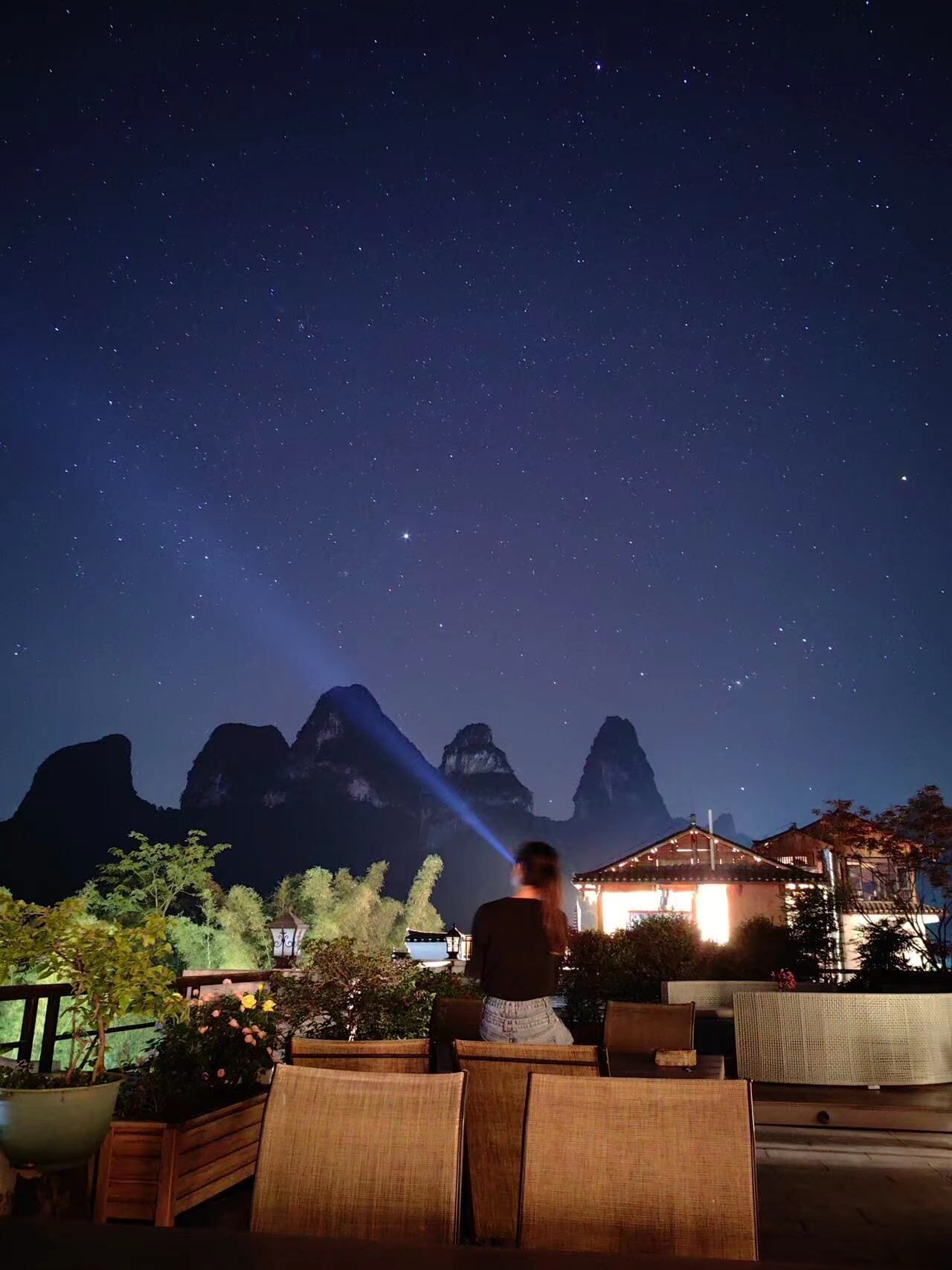 Une personne se tient dans un espace extérieur aménagé en coin salon, avec une montagne en arrière-plan et un ciel étoilé.